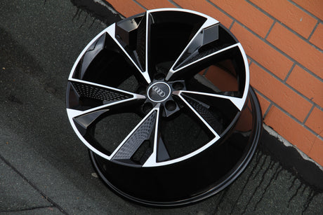 An Audi car wheel rim rests against a brick wall. It features a sleek black and silver geometric design with the Audi logo at the center. The ground is textured.