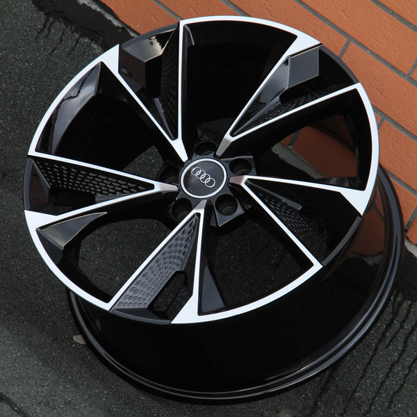 A glossy black and silver alloy wheel featuring the Audi logo rests against a brick wall on a textured pavement.