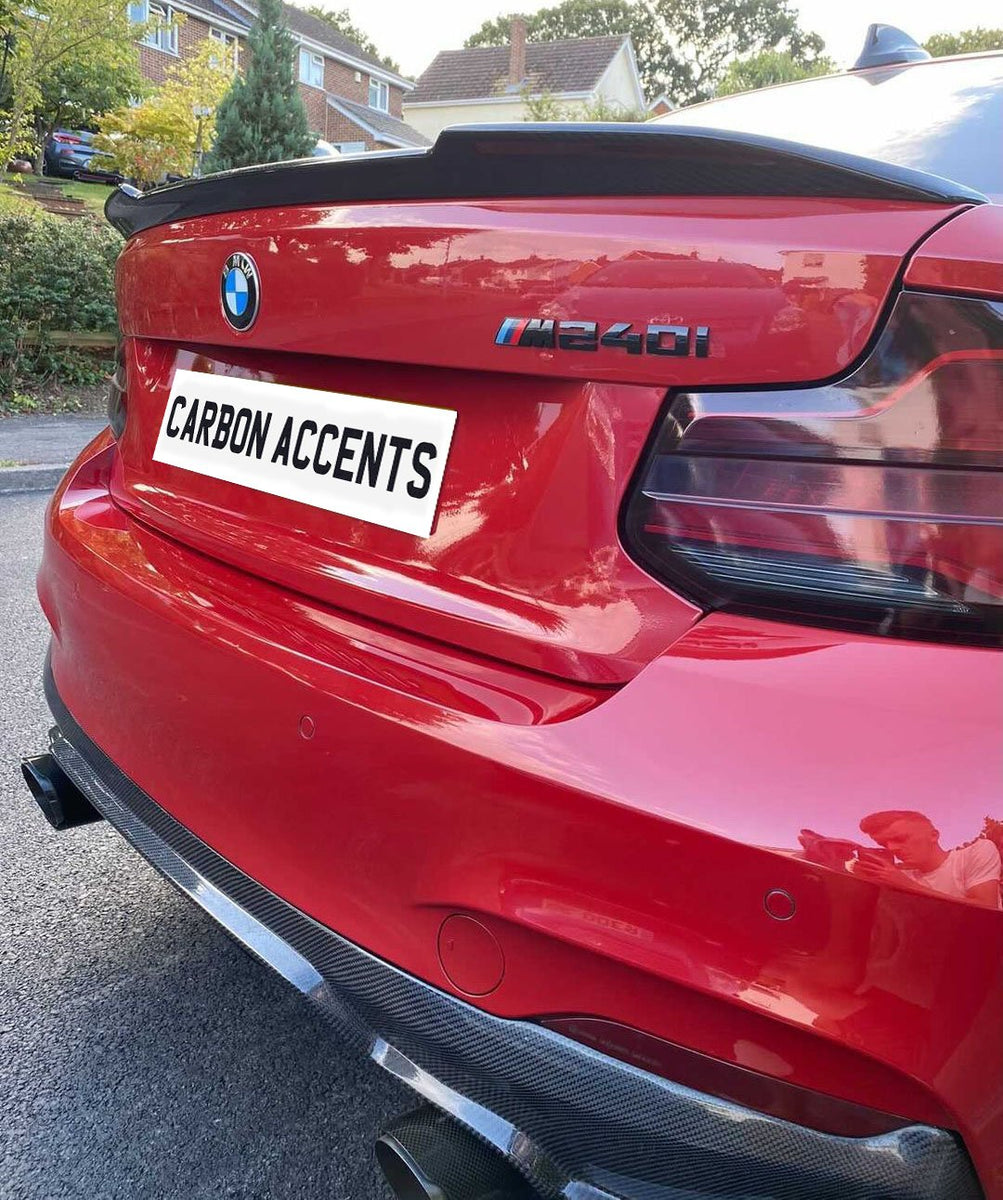 Red BMW M240i parked on a residential street, showcasing carbon fiber accents and dual exhaust. License plate reads "CARBON ACCENTS." Houses and greenery form the background.