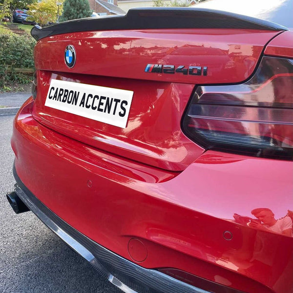 Red BMW M240i parked on a residential street, showcasing carbon fiber accents and dual exhaust. License plate reads "CARBON ACCENTS." Houses and greenery form the background.