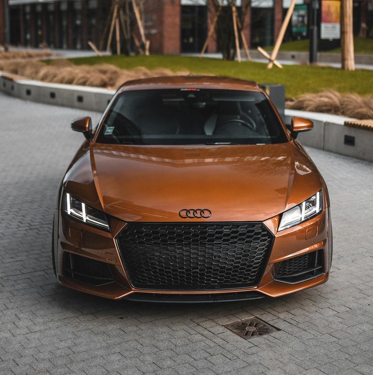 A bronze Audi sports car is parked on a brick-paved driveway surrounded by landscaped grass and modern buildings. The car's headlights are on, and its grille is prominently visible.