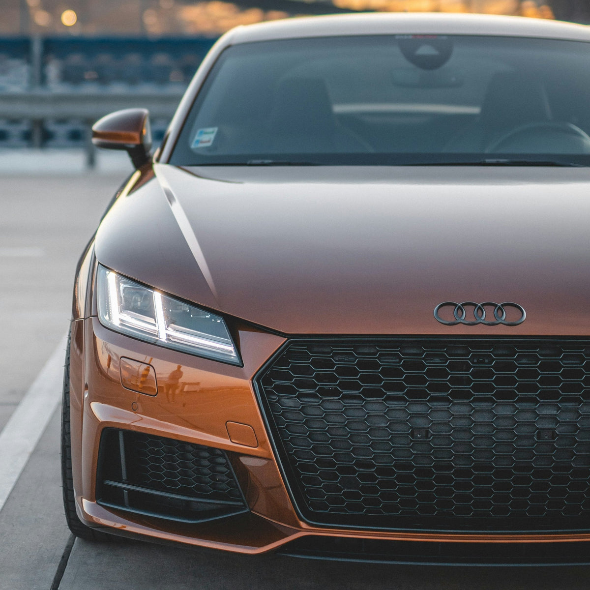 A bronze Audi sports car sits parked, its sleek front grille and headlights visible. It rests on a concrete pavement with metal barriers and a sunset sky in the background.
