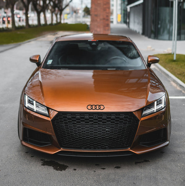A metallic bronze Audi sports car is parked on a street near modern buildings and a grassy area. Its sleek design features angular headlights and a prominent grille with the Audi logo.