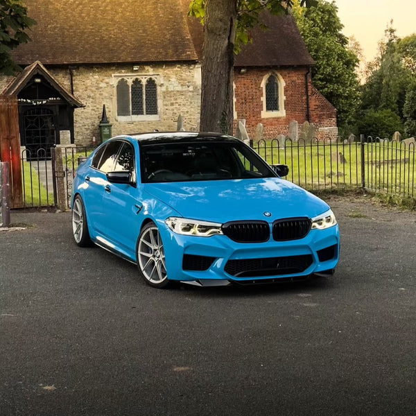 A blue car is parked on a paved area near a churchyard with a historic stone building and gravestones, surrounded by a metal fence and trees.