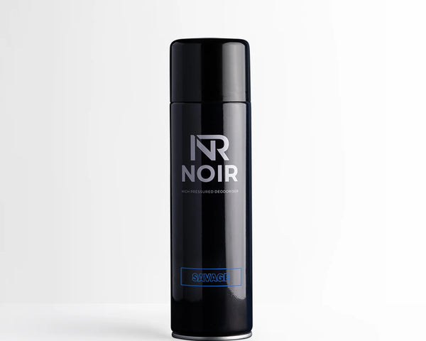 A black cylindrical deodorant can stands upright on a white surface. Text on the can reads "NR NOIR HIGH PRESSURED DEODORISER SAVAGE," set in a minimalistic and bright environment.