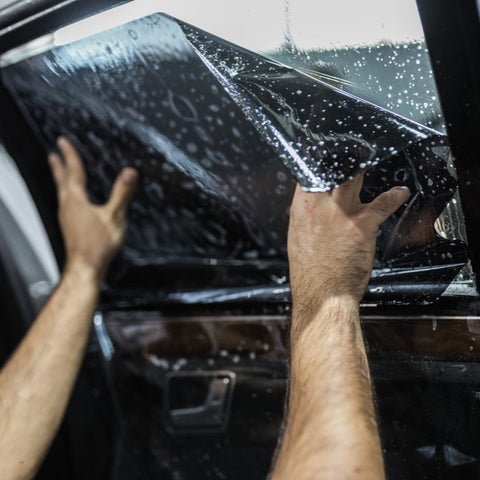 Hands apply dark film to a car window speckled with water droplets, inside a vehicle, creating a privacy tint.
