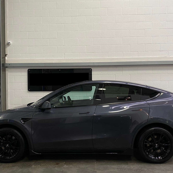 A sleek, gray car is parked indoors. It faces left, next to a metal shutter with a white brick wall in the background, creating an industrial setting.