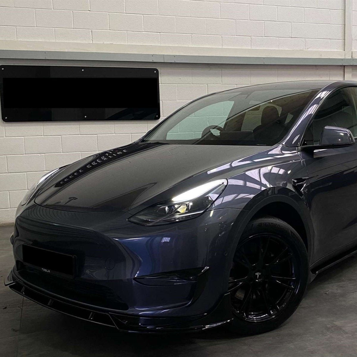A sleek, dark-colored car is parked indoors near a white brick wall. Its headlights and aerodynamic design are prominent, with a reflective surface displaying overhead lighting. No text is visible.
