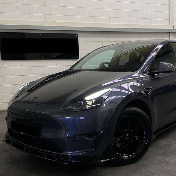 A sleek, dark-colored car is parked indoors near a white brick wall. Its headlights and aerodynamic design are prominent, with a reflective surface displaying overhead lighting. No text is visible.