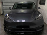 The car, a dark gray Tesla, is parked facing forward inside a garage with a closed, corrugated metal door and white brick walls. The front license plate reads 