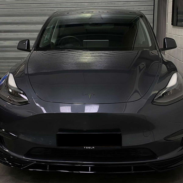The car, a dark gray Tesla, is parked facing forward inside a garage with a closed, corrugated metal door and white brick walls. The front license plate reads "TESLA."