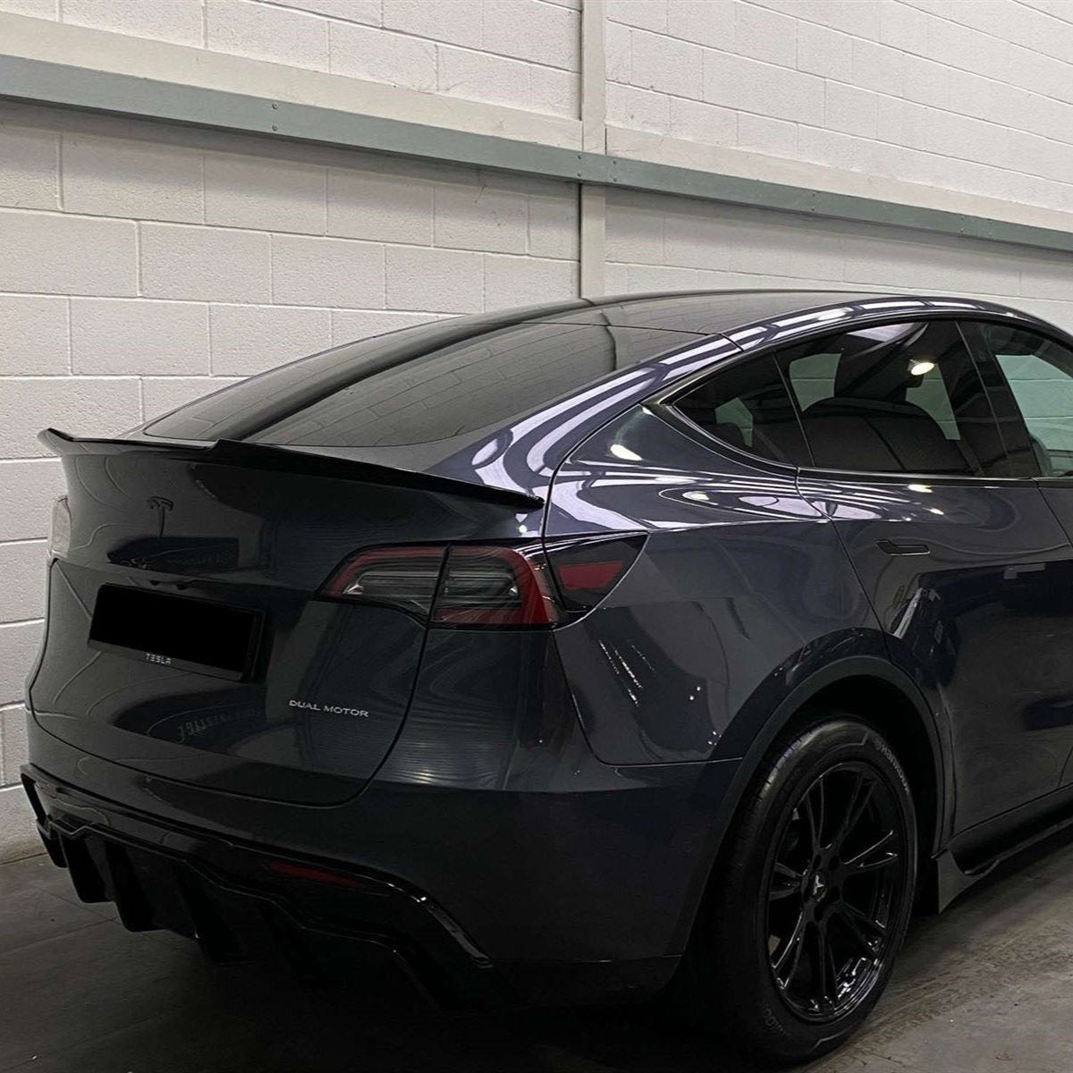 A dark-colored car with a spoiler is parked indoors against a white brick wall. Text on the car reads "DUAL MOTOR." Reflections are visible on its glossy surface.