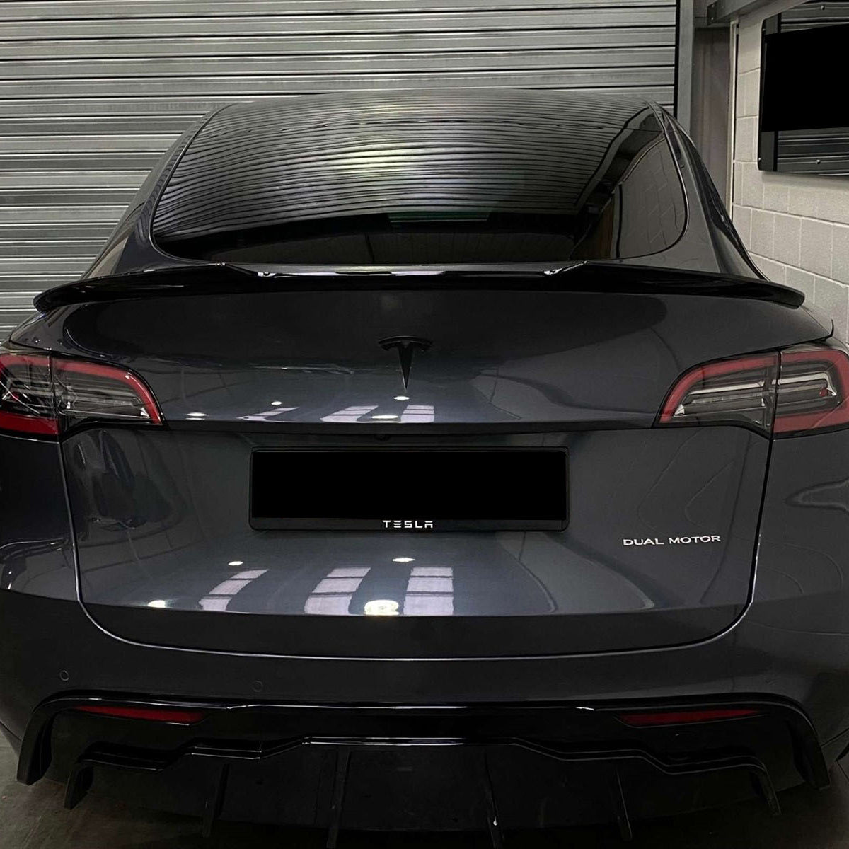 A black Tesla car is parked inside a garage. The back features "TESLA" and "DUAL MOTOR" text, with a closed metallic shutter and a wall-mounted screen in the background.