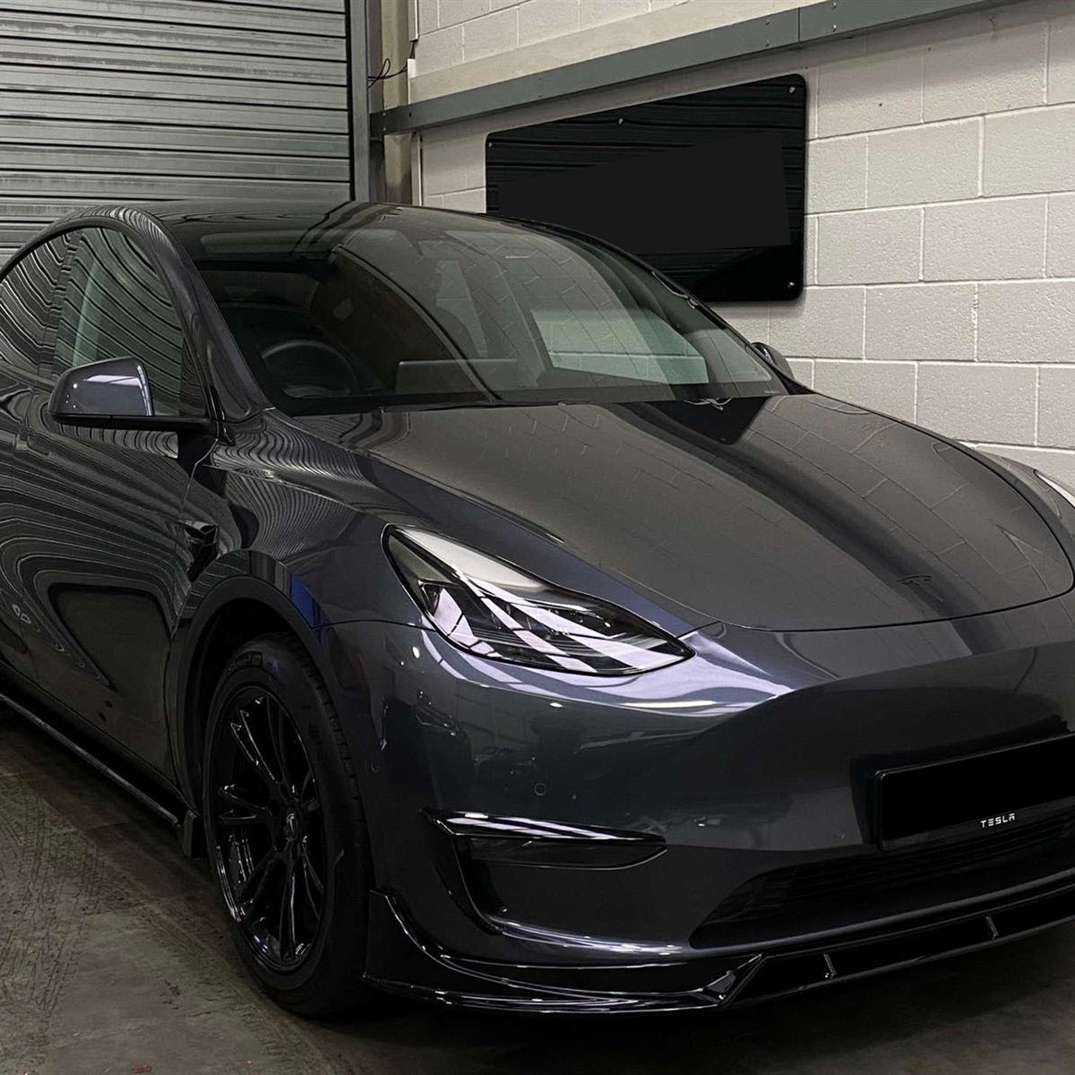 A sleek, gray car sits stationary, facing front-left, in an indoor garage with roller doors and white walls. "TESLA" is visible on the front bumper.