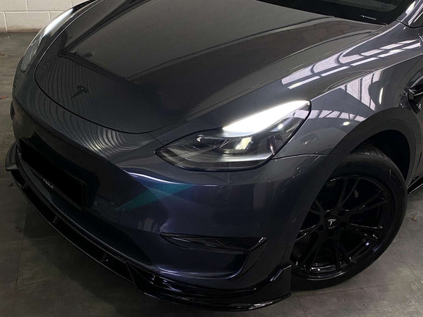 A dark-colored Tesla car with illuminated headlights is parked indoors. The focus is on the front and side, displaying its sleek design and black wheels in dim lighting.