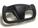 A sleek Tesla yoke steering wheel, featuring minimalistic controls, rests on a clear stand. It is black with 