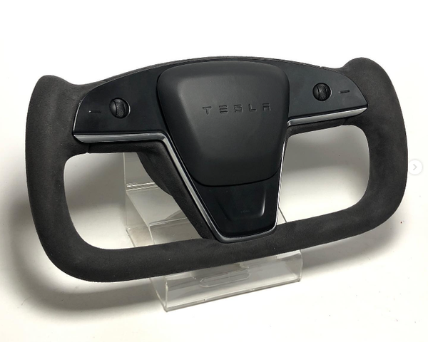 A sleek Tesla yoke steering wheel, featuring minimalistic controls, rests on a clear stand. It is black with "TESLA" inscribed at the center, set against a plain white background.