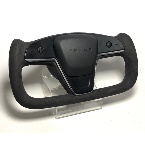 A sleek Tesla yoke steering wheel, featuring minimalistic controls, rests on a clear stand. It is black with "TESLA" inscribed at the center, set against a plain white background.