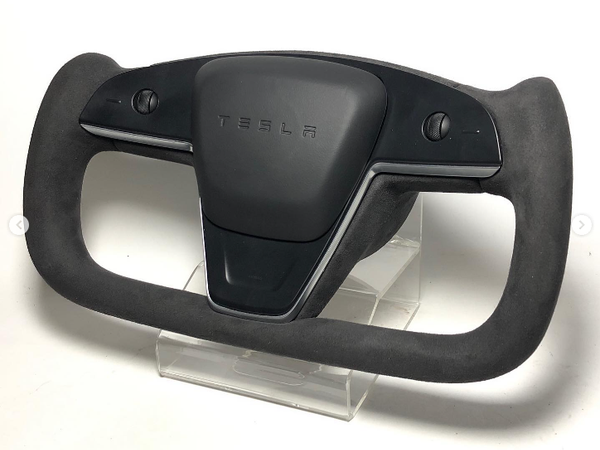 A black Tesla steering wheel with a yoke design, resting on a clear stand. The center features the Tesla logo embossed, and buttons are visible on either side.