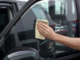 A hand cleans a car's side window using a beige cloth, in a garage with other vehicles and large windows in the background.