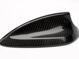 A shark fin-shaped car antenna made from carbon fiber, resting on a white surface. It features a glossy finish and a sleek, aerodynamic design.