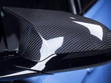 A carbon fiber car side mirror reflects light, highlighting its texture. It is attached to a sleek, glossy vehicle exterior with a blue background, showcasing a modern automotive design.