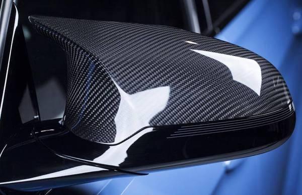 A carbon fiber car side mirror reflects light, highlighting its texture. It is attached to a sleek, glossy vehicle exterior with a blue background, showcasing a modern automotive design.