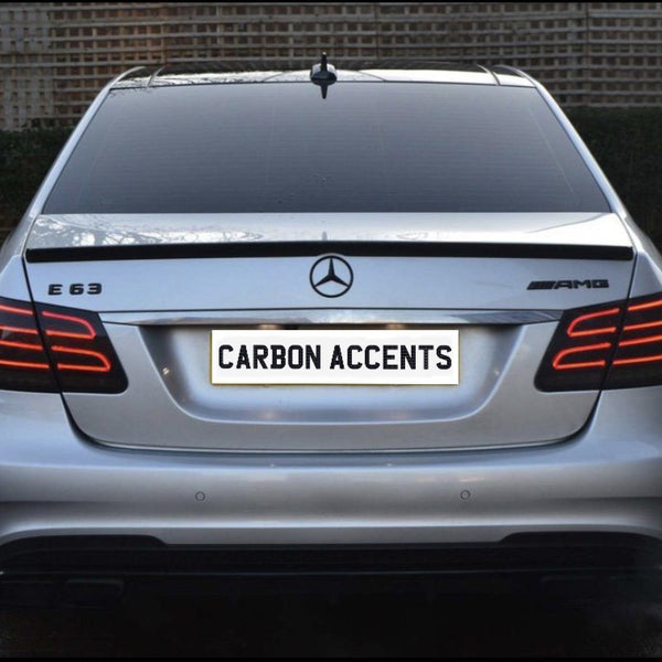 The image shows the rear of a silver Mercedes-Benz E63 AMG with illuminated tail lights, parked on a brick driveway. The license plate reads "CARBON ACCENTS." The background features a wooden fence.