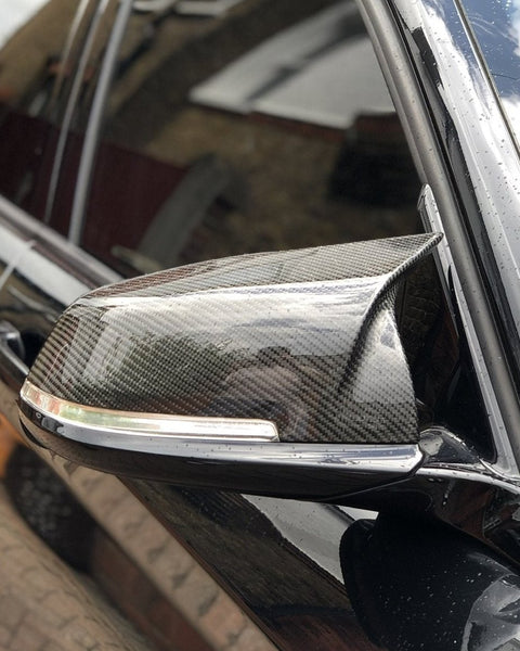 A carbon fiber car side mirror with integrated indicator light on a shiny black vehicle, parked near a brick wall reflecting the cloudy sky.