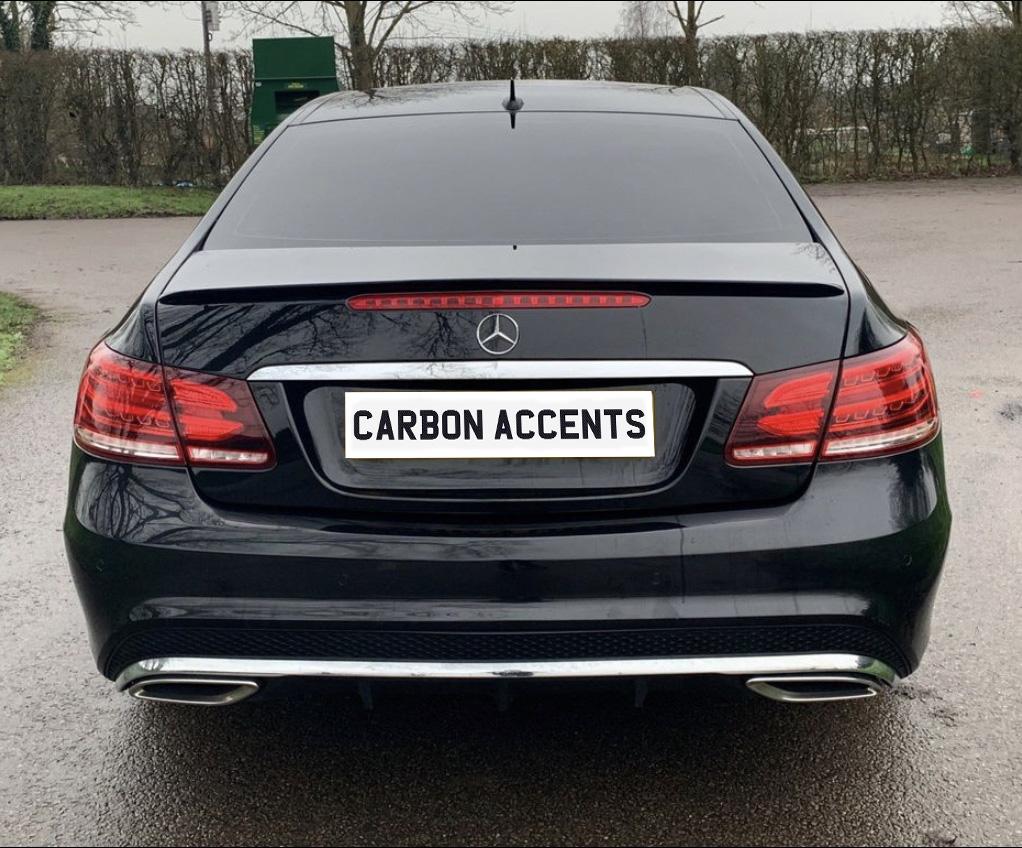 Car with black paint and a "CARBON ACCENTS" license plate remains stationary in a parking area. Background includes leafless trees and a green container.