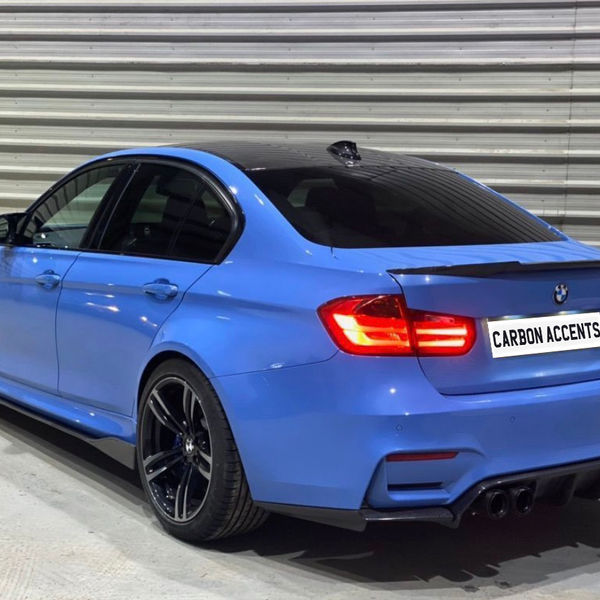 A blue BMW sedan is parked indoors, featuring black wheels and bright taillights. The rear license plate reads "CARBON ACCENTS," against a backdrop of metallic, corrugated walls.