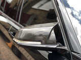 A carbon fiber side mirror is attached to a black car, reflecting buildings and sky, parked on cobblestone pavement.