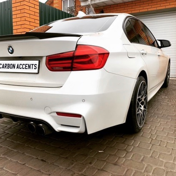 A white BMW sedan is parked on a cobblestone driveway, adjacent to a brick building with a green fence. The license plate reads "CARBON ACCENTS."