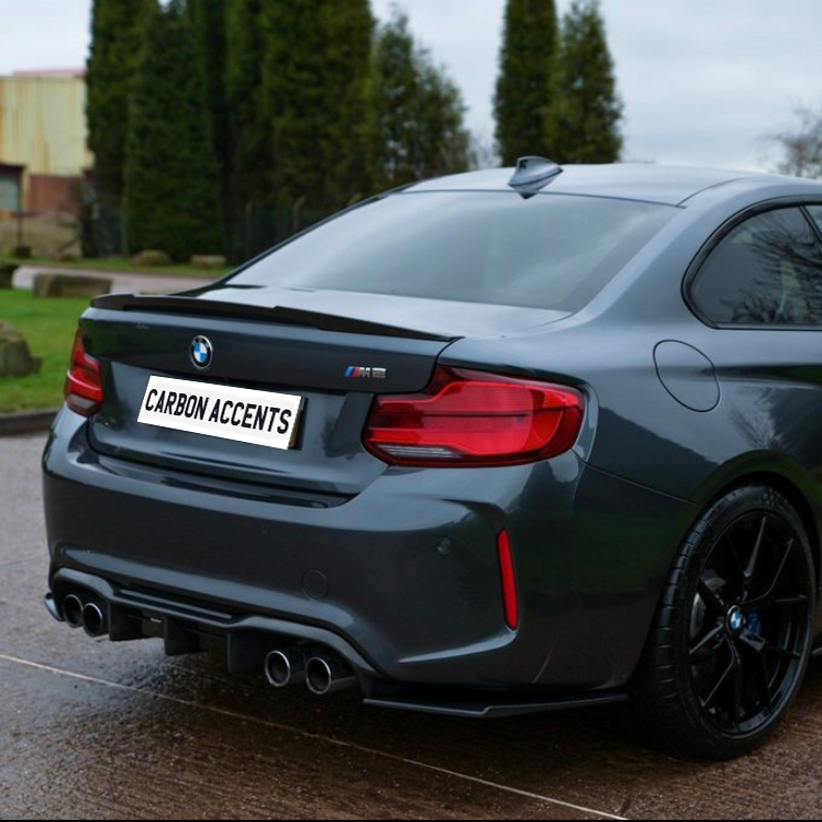 A gray sports car is parked on a wet road beside green grass and tall trees. The license plate reads "CARBON ACCENTS," and it has four visible exhaust pipes.