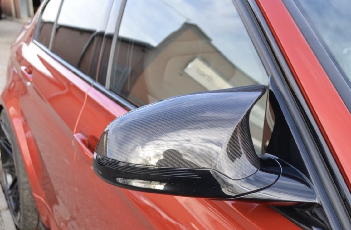 A car's side mirror, featuring a carbon fiber texture, reflects a cloudy sky. The vibrant red vehicle is parked in an outdoor setting, with a building visible in the background.