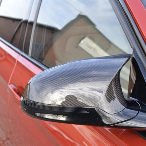 A car's side mirror, featuring a carbon fiber texture, reflects a cloudy sky. The vibrant red vehicle is parked in an outdoor setting, with a building visible in the background.