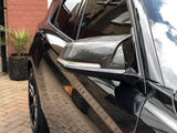 A shiny black car with carbon fiber side mirrors is parked on a brick driveway. The reflection shows nearby buildings and sky. A plant and brick wall are visible nearby.