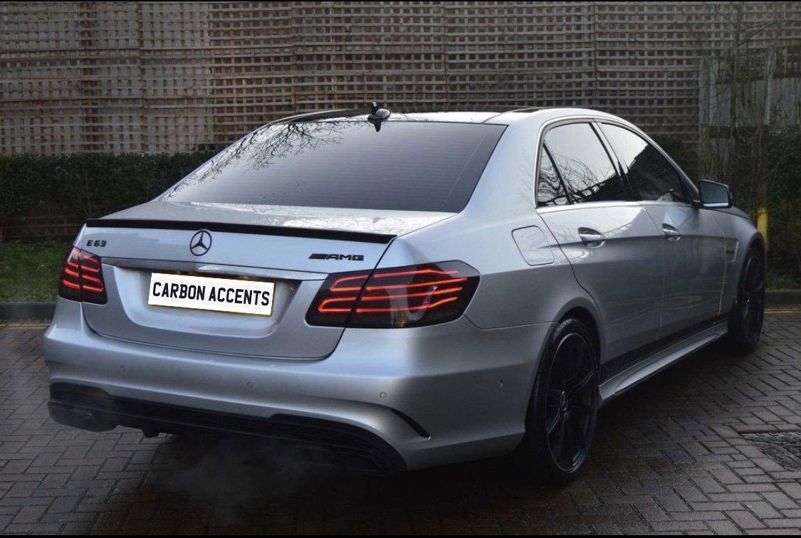 A silver Mercedes AMG E63 is parked on a brick surface, emitting exhaust vapor. The license plate reads "CARBON ACCENTS," with wooden fencing and greenery in the background.
