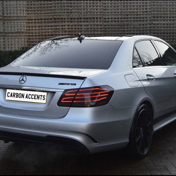 A silver Mercedes AMG E63 is parked on a brick surface, emitting exhaust vapor. The license plate reads "CARBON ACCENTS," with wooden fencing and greenery in the background.