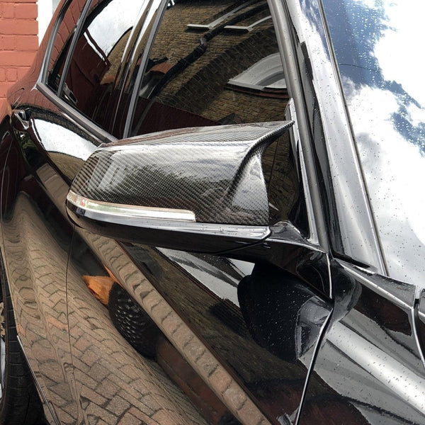 A carbon fiber car side mirror reflects the brick building and cloudy sky, with water droplets visible on the black vehicle in a driveway setting.