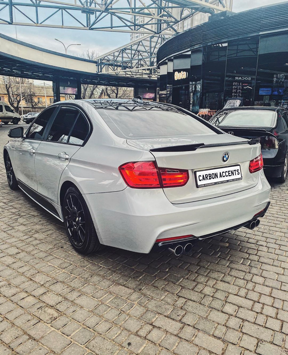 A silver BMW sedan is parked on a cobblestone area near a storefront labeled "Buta" with a "CARBON ACCENTS" license plate, surrounded by other vehicles and structures.