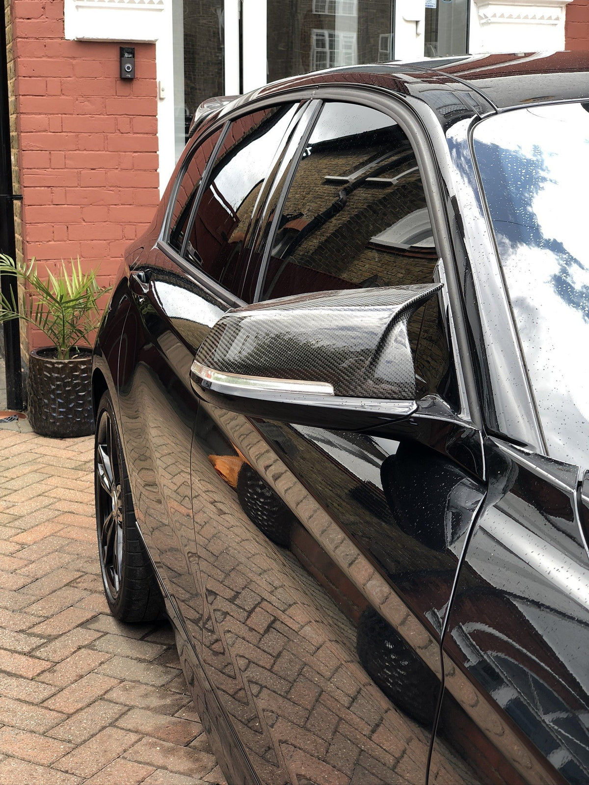 A black car with tinted windows and carbon fiber side mirror is parked on a brick driveway. Its reflective surface displays a brick wall and glass doors in an urban setting.
