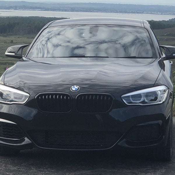 A black BMW car is parked on a road, facing forward, with rolling hills and a body of water in the distant background.