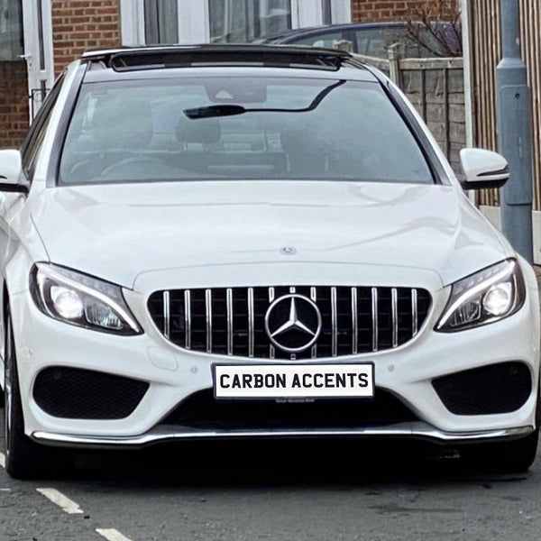 Car parked on a residential street, with LED headlights on. It's a white Mercedes-Benz with distinctive grille and license plate reading "CARBON ACCENTS." Bricks and lampposts are visible nearby.