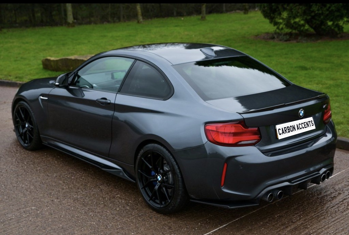 A dark gray sportscar with tinted windows is parked on a wet road. The license plate reads "CARBON ACCENTS," and lush green grass borders the scene.