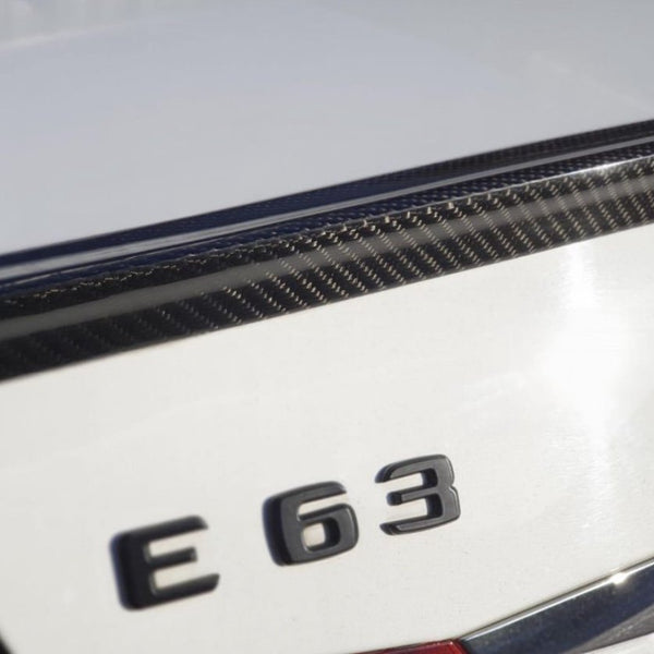 The image shows the rear emblem "E 63" on a white car, featuring a carbon fiber detail on the trunk lid in bright sunlight.