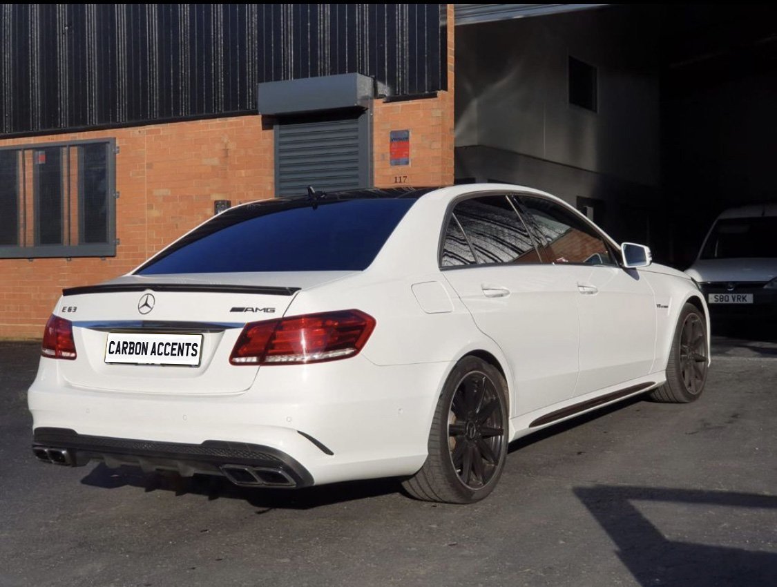 A white Mercedes-Benz E63 AMG is parked on a paved area outside an industrial building with orange brick walls. The text "CARBON ACCENTS" is on the license plate.