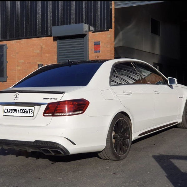 A white Mercedes-Benz E63 AMG is parked on a paved area outside an industrial building with orange brick walls. The text "CARBON ACCENTS" is on the license plate.