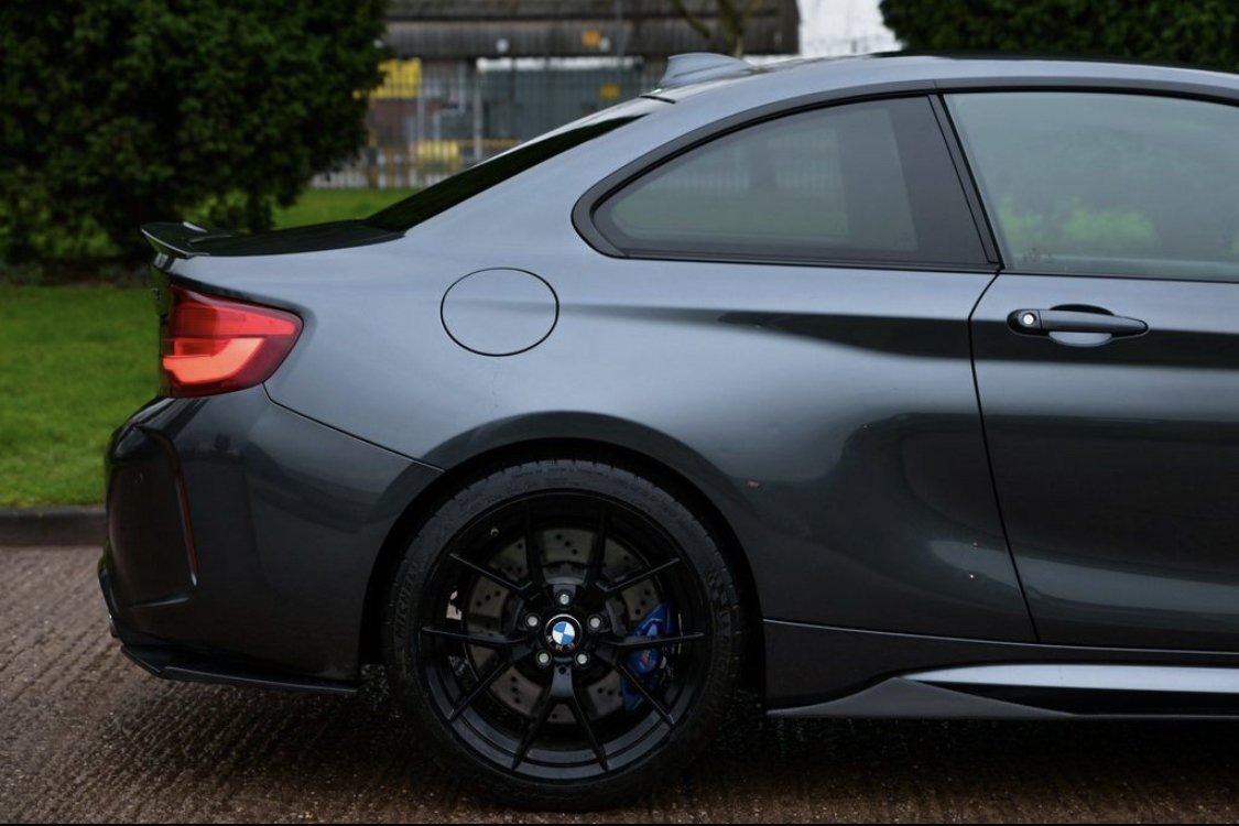 A sleek gray car is parked on a paved surface, showcasing its rear side and black alloy wheel, with green grass and a fence in the background.