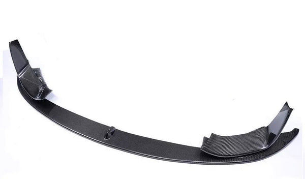 Carbon fiber car bumper lip sits on a plain white surface, showcasing its curved design and textured finish, with raised ends for aerodynamic improvement.